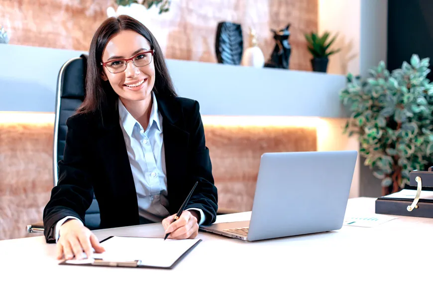 Junge Frau mit braunen langen Haaren und Brille, schick gekleidet sitzt am Schreibtisch mit ihrem Laptop und notiert sich etwas.